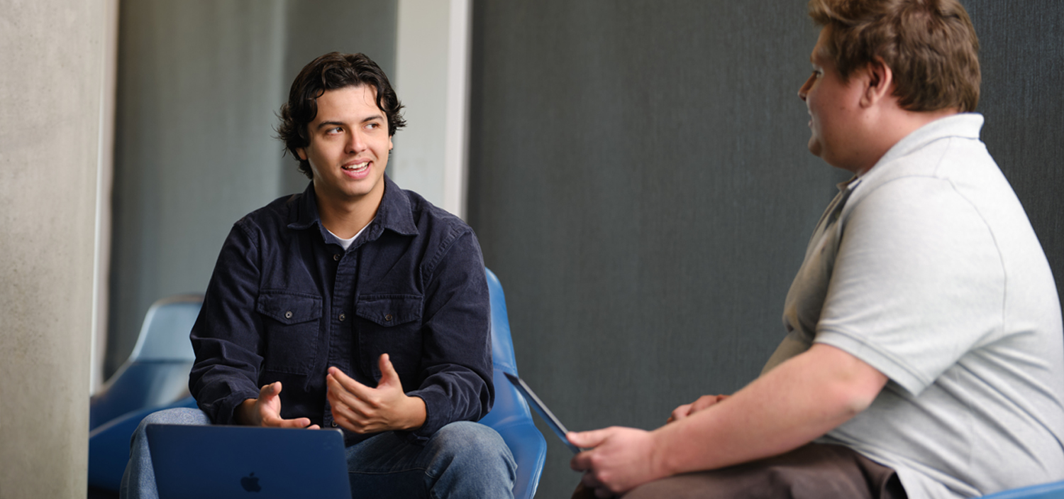 Two students sitting in armchairs looking at each other with laptops on a coffee table.